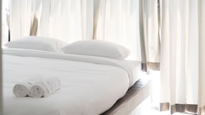 Una cama bien hecha con sábanas blancas y almohadas, una toalla enrollada y cortinas translúcidas entreabiertas.