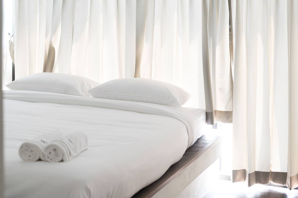 A neatly made bed with white linens and pillows, a rolled towel, and sheer curtains partially drawn.