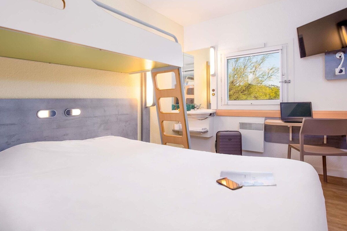A hotel room with a bunk bed, a desk with a laptop, a chair, a window with a view, and a TV mounted on the wall.