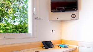 A room with a window, a flat-screen TV mounted on the wall, a desk with a tablet and a smartphone, and a small stool. - ibis budget Fréjus Saint-Raphaël Capitou A8 (Fréjus)
