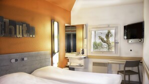 A hotel room with two beds, a desk, a chair, a TV, a bathroom, and a window with a view of trees.