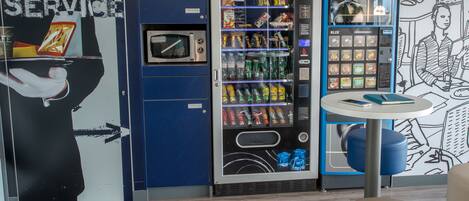 Een automaat met diverse snacks en dranken, een magnetron en een klein tafeltje met een stoel.