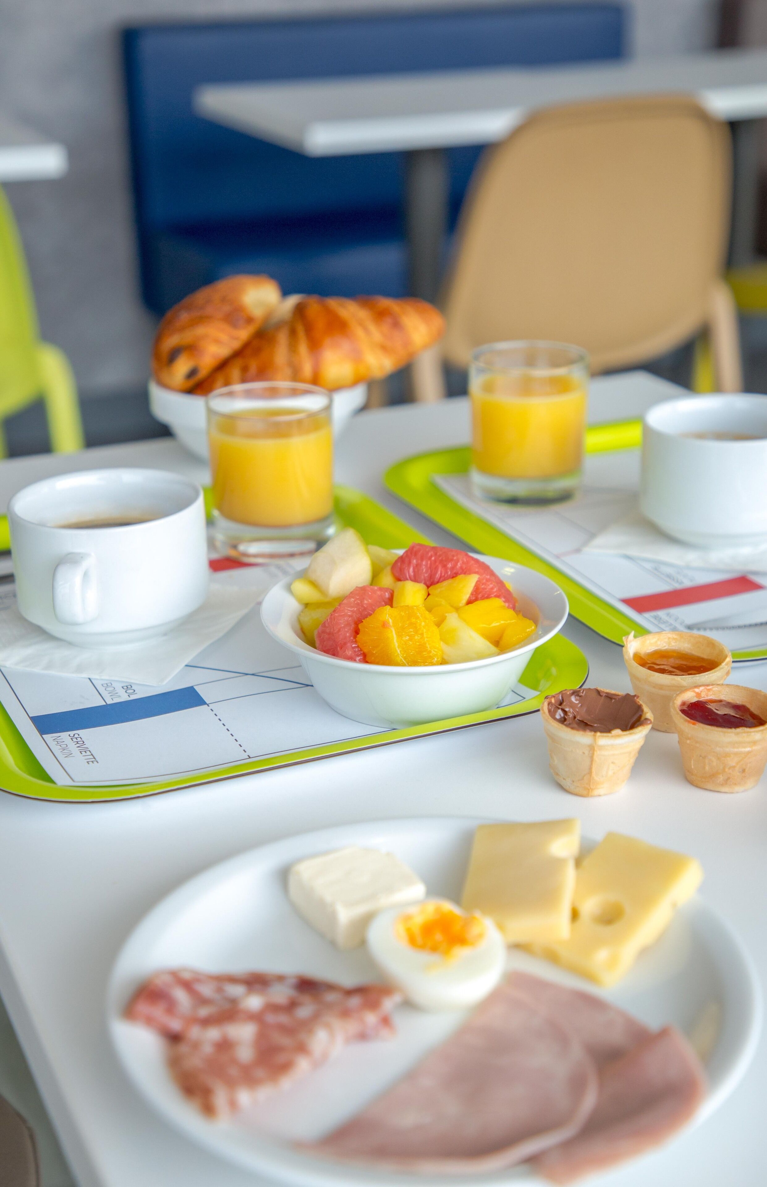 Una mesa de desayuno con cruasanes, jugo de naranja, un bol de fruta, huevos, queso y jamón sobre una bandeja.