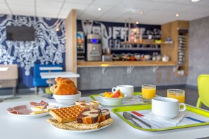 A breakfast setting with waffles, eggs, and fruit, and a coffee bar in the background. - ibis budget Fréjus Saint-Raphaël Centre et Plage (Fréjus)