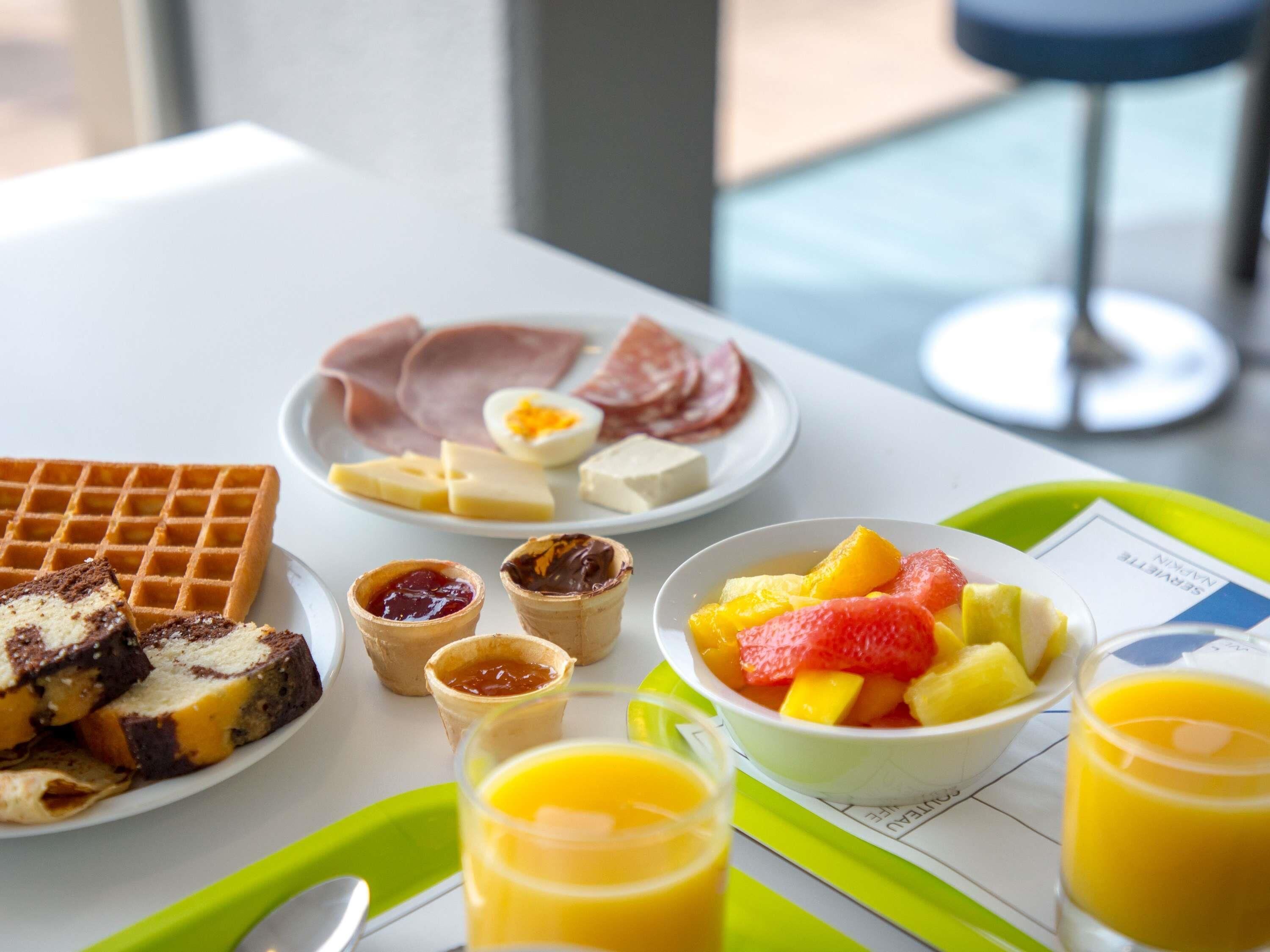 Un plateau de petit-déjeuner avec des gaufres, du fromage, du jambon, des œufs, des fruits et du jus d’orange.