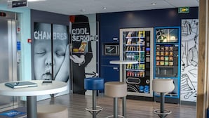 A modern interior with a vending machine, seating area, and a wall with a large advertisement. - ibis budget Fréjus Saint-Raphaël Centre et Plage (Fréjus)