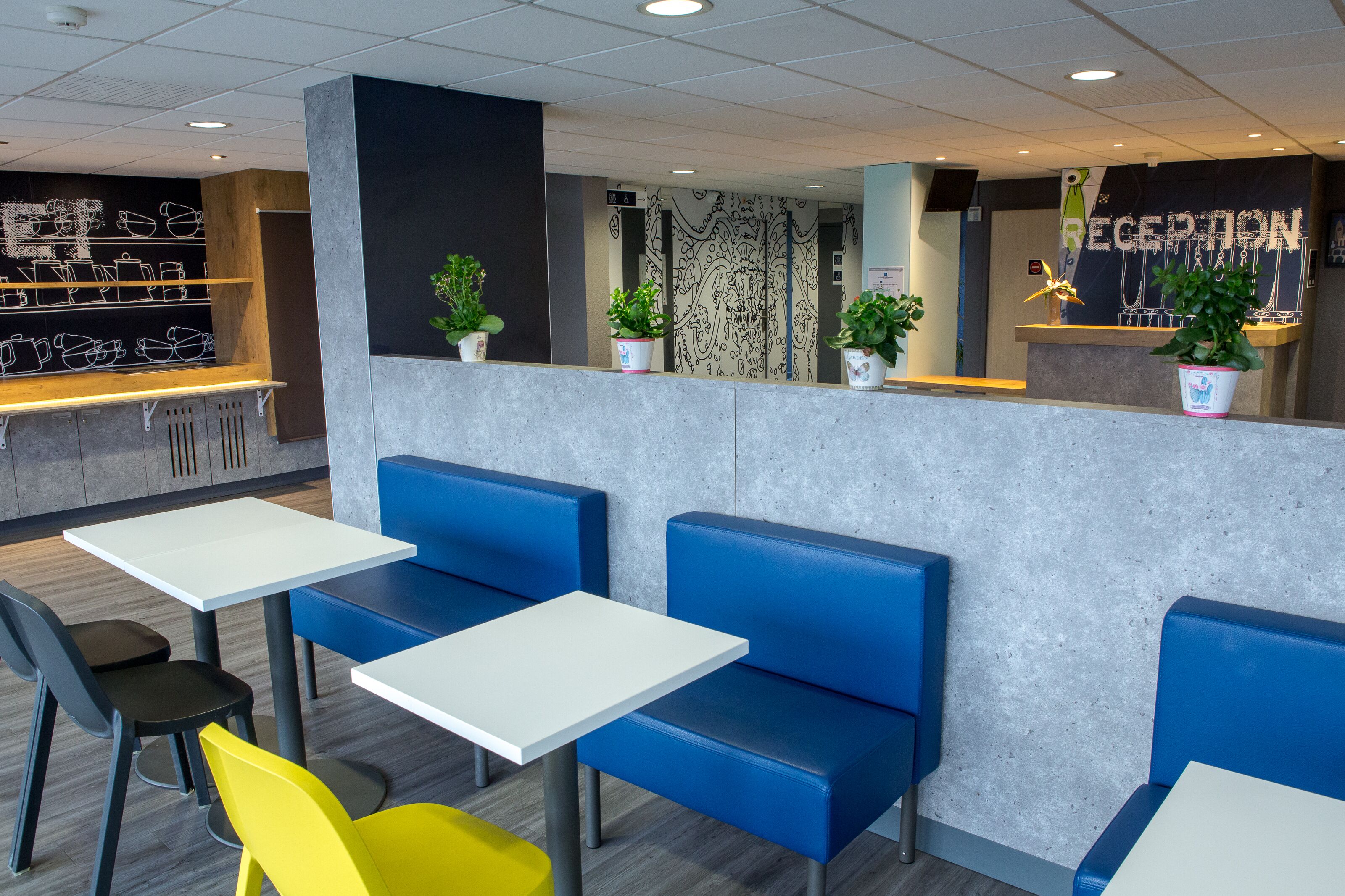 Un espace d’accueil moderne comprenant un comptoir de réception, un coin salon avec des banquettes bleues et un mur décoré sur le thème du café.