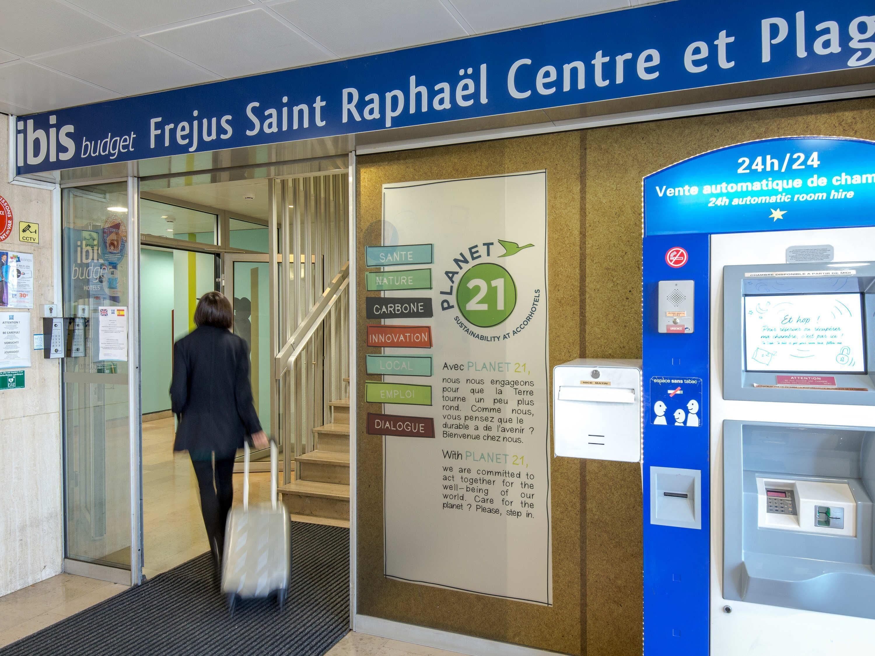 L’entrée d’un hôtel, avec une enseigne indiquant « ibis budget Fréjus Saint-Raphaël Centre et Plage » et un distributeur automatique de clés pour les chambres, accessible 24 heures sur 24.