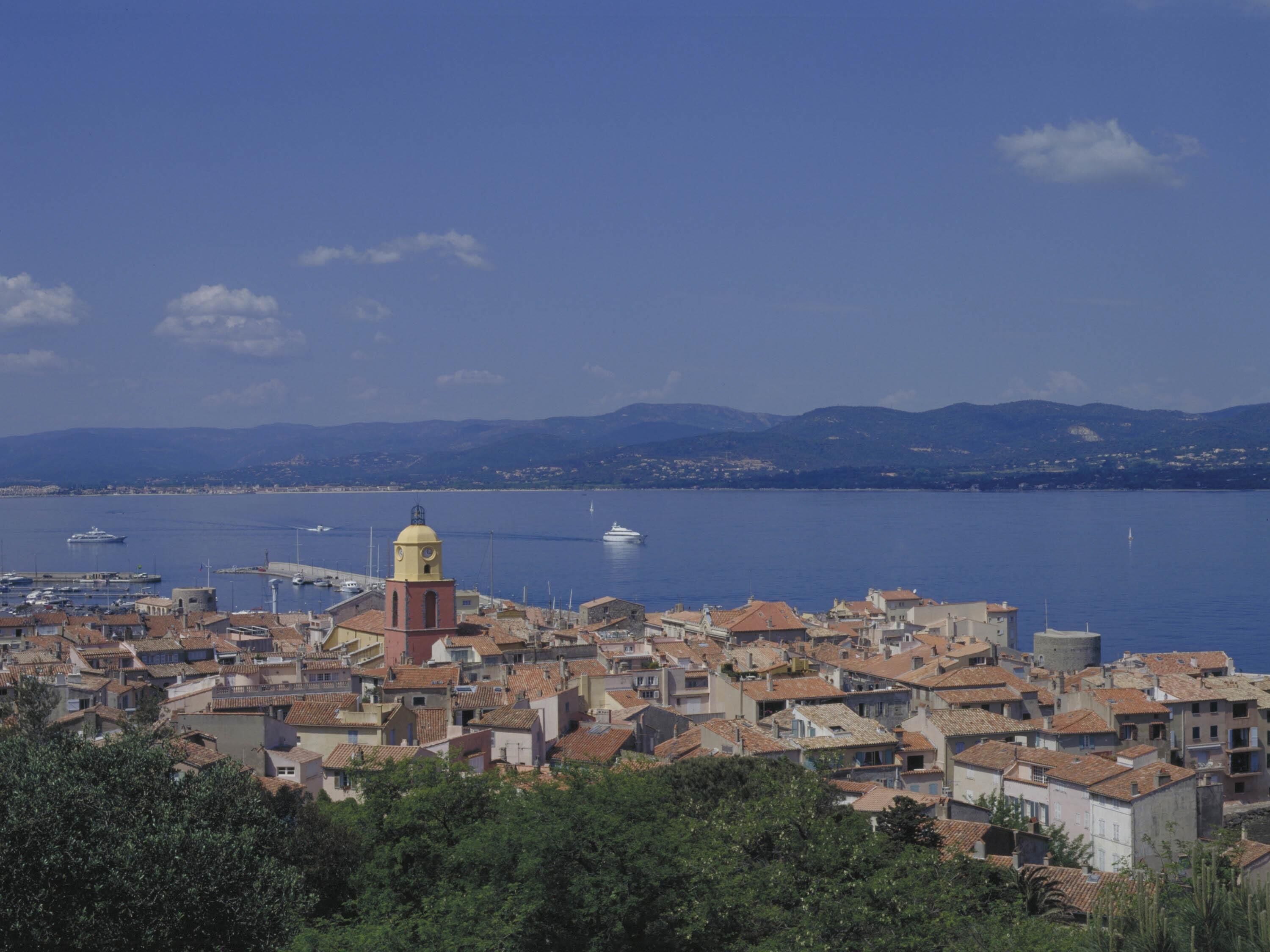 A coastal town with a prominent yellow tower, dense buildings with red-tiled roofs, and a marina with boats.