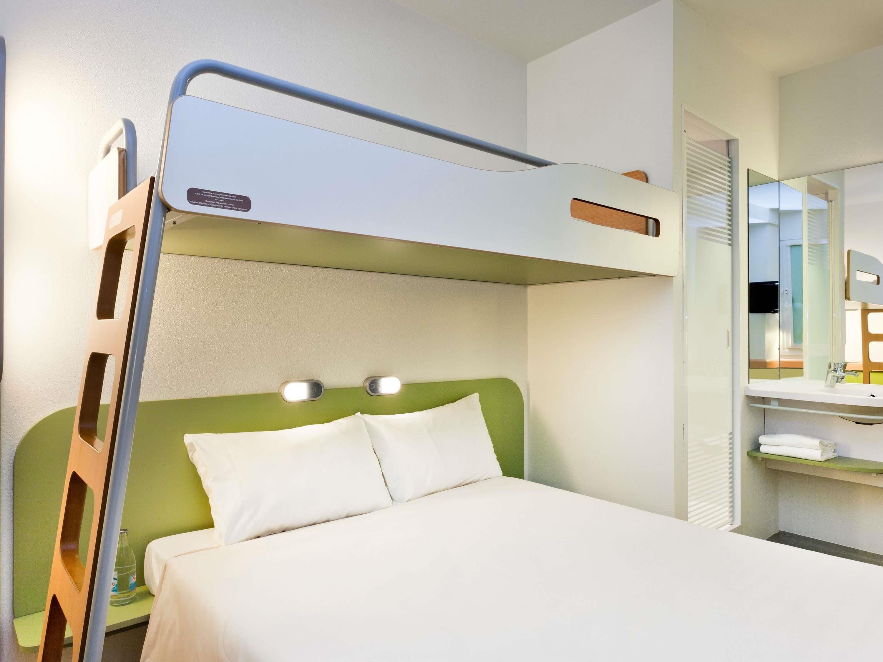 Una cama litera con cabecera verde, una mesita de noche con lámpara y un espejo sobre la cama.