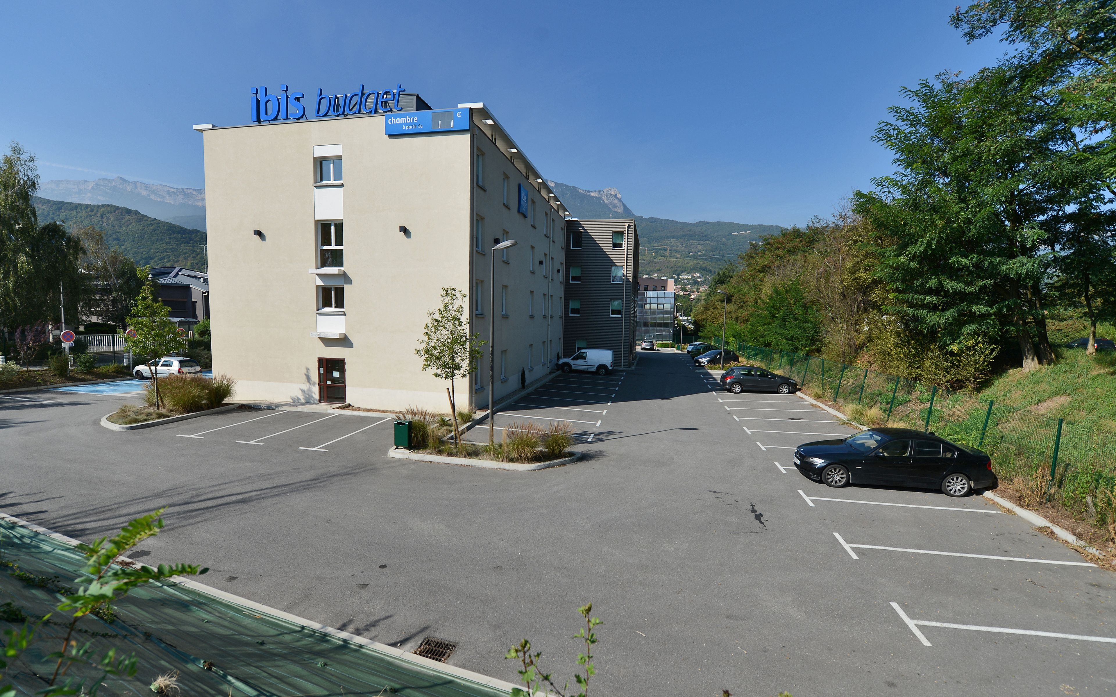 Un hotel llamado "ibis budget" con un estacionamiento, una camioneta blanca y una montaña al fondo.