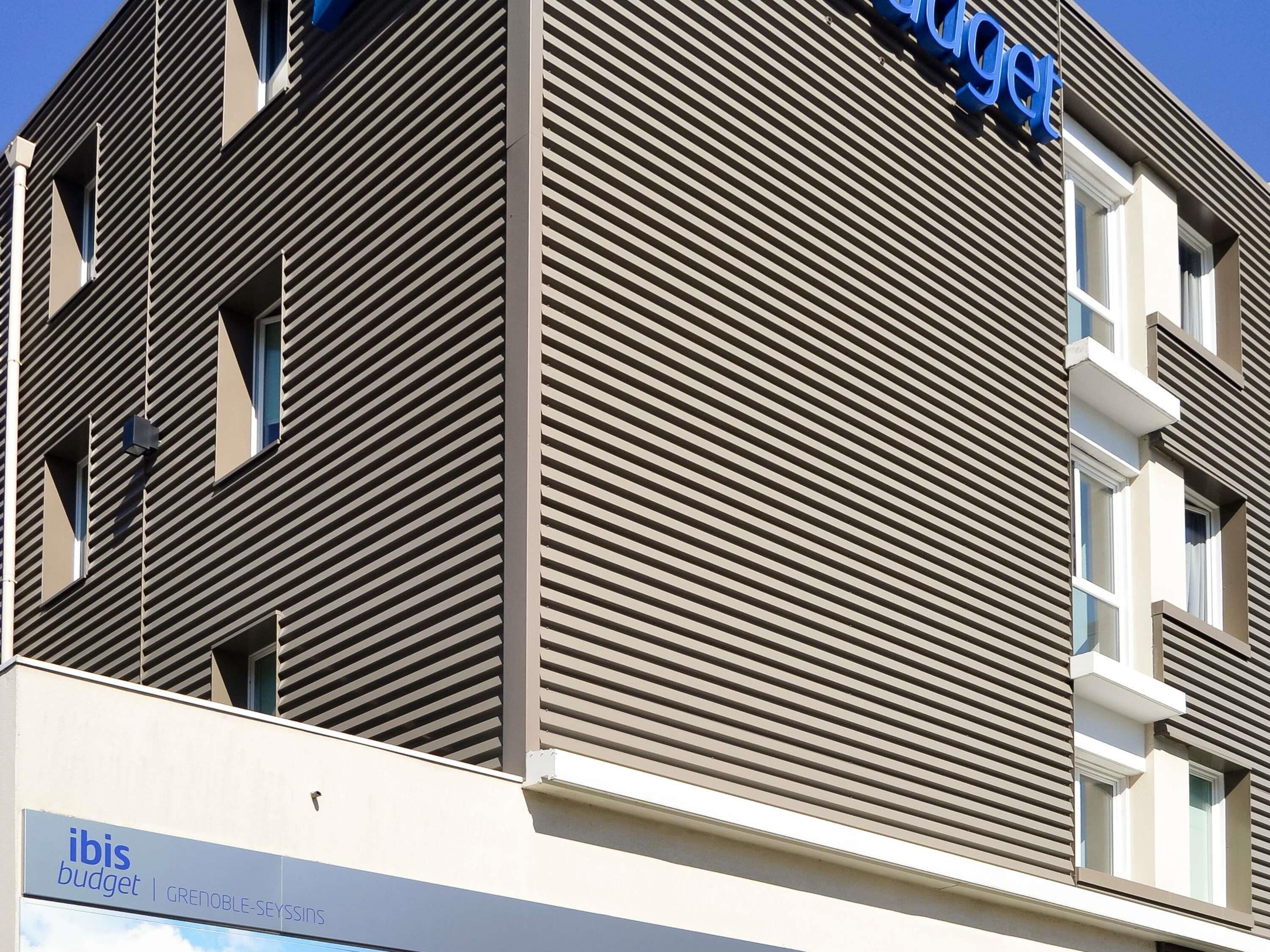 A modern building with horizontal slats, a visible sign for 'ibis budget Grenoble-Seyssis,' and a clear blue sky.