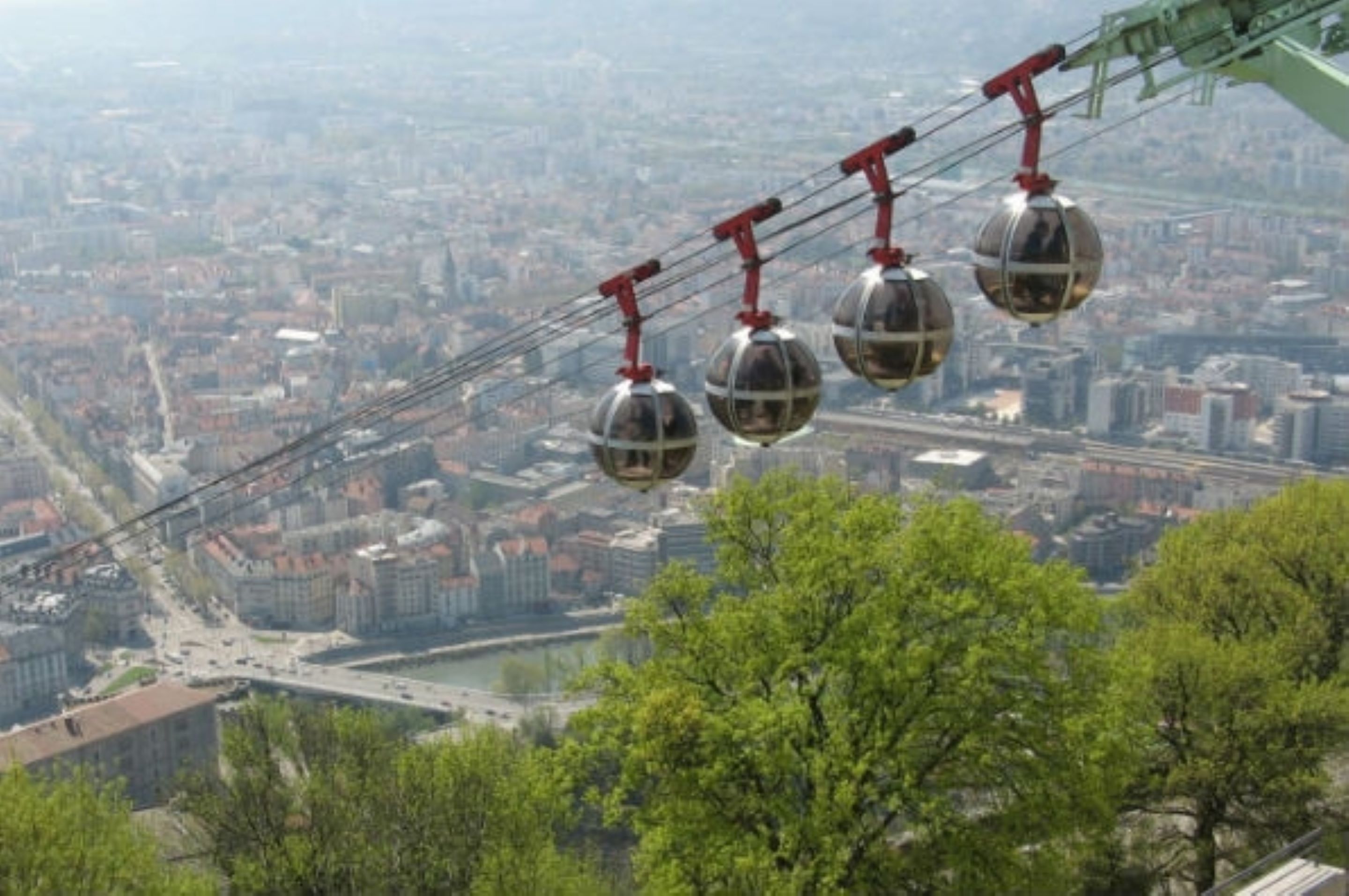 Un téléphérique composé de cabines sphériques suspendues à des câbles rouges, offrant une vue panoramique sur une ville avec ses bâtiments et une rivière.