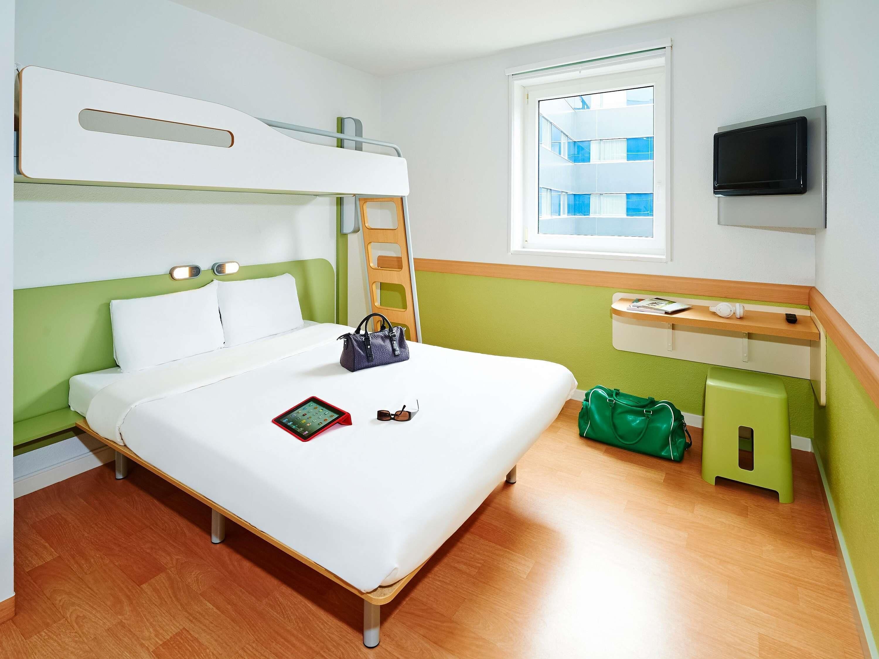 Habitación de hotel con literas, una mesita de noche con televisor, una ventana con vistas a edificios y un bolso verde en el suelo.
