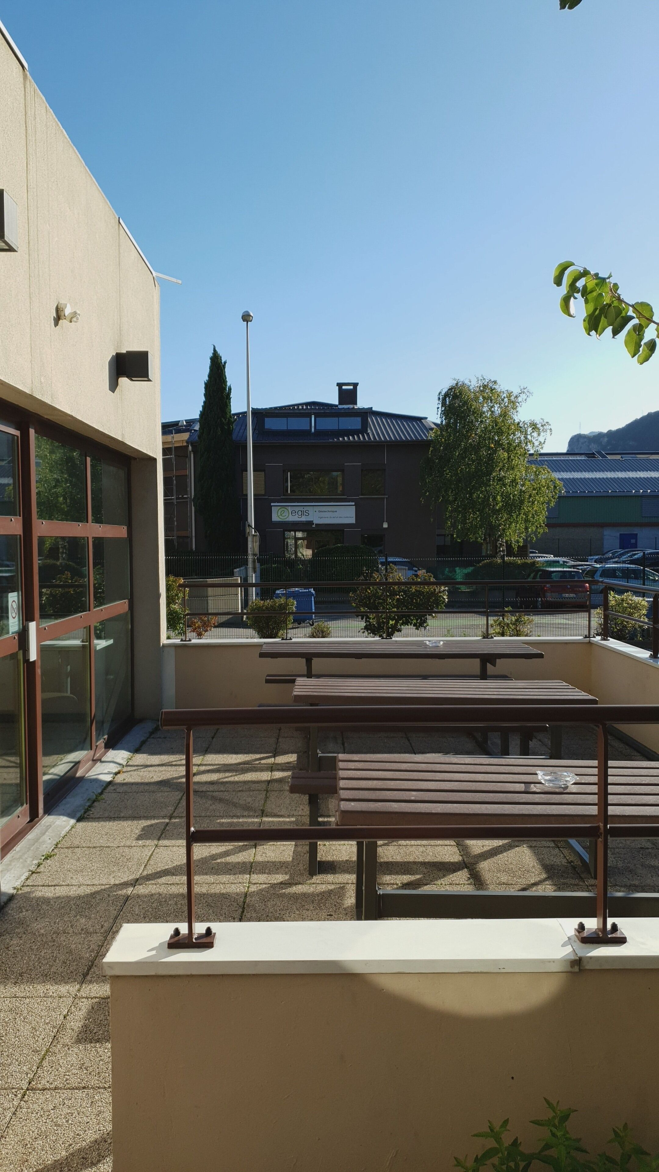 An outdoor seating area with benches and a table, surrounded by a low wall and a building with large windows.