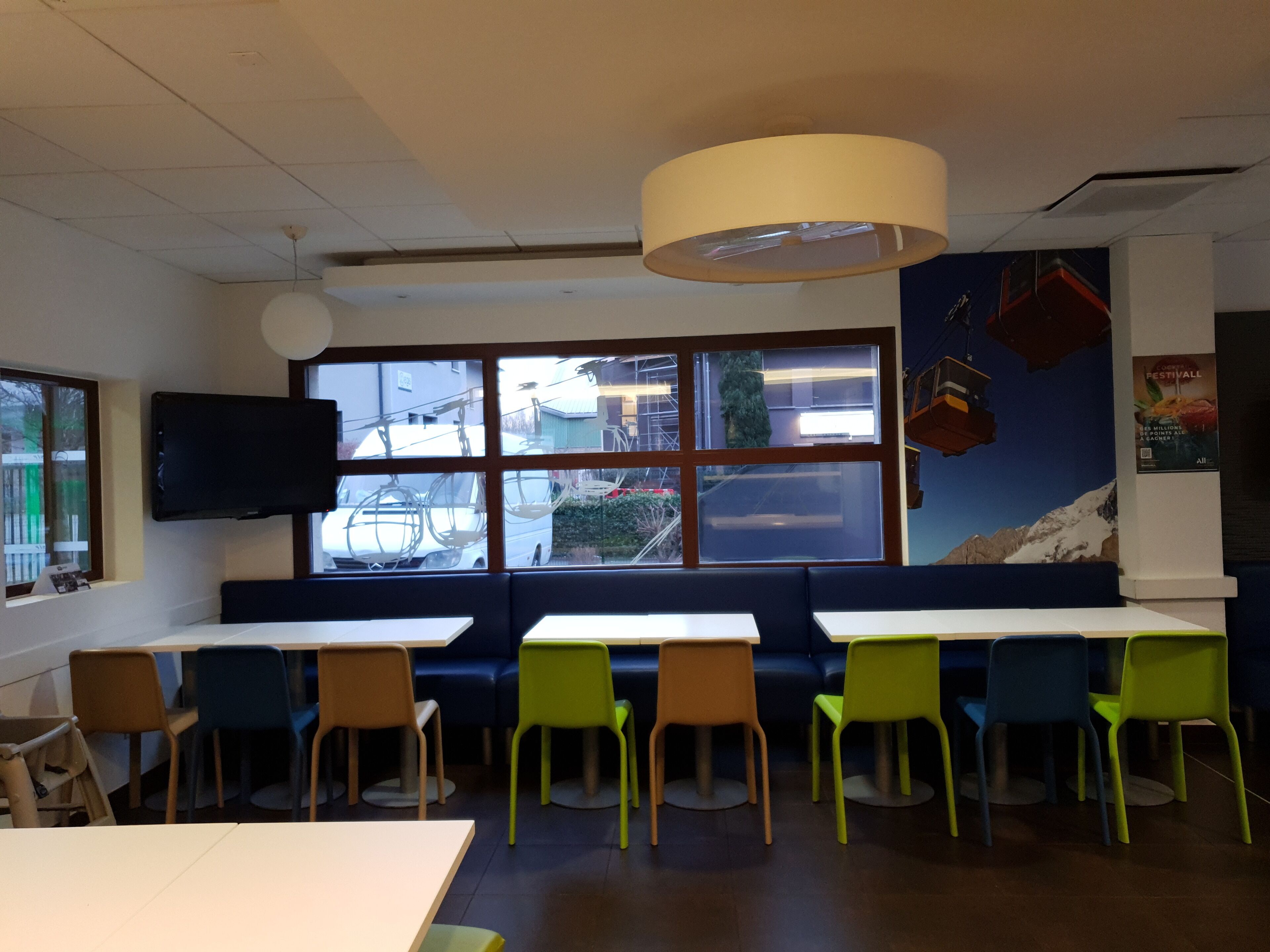 Un interior de restaurante con mesas y sillas, un ventanal con vistas a la calle y un televisor montado.