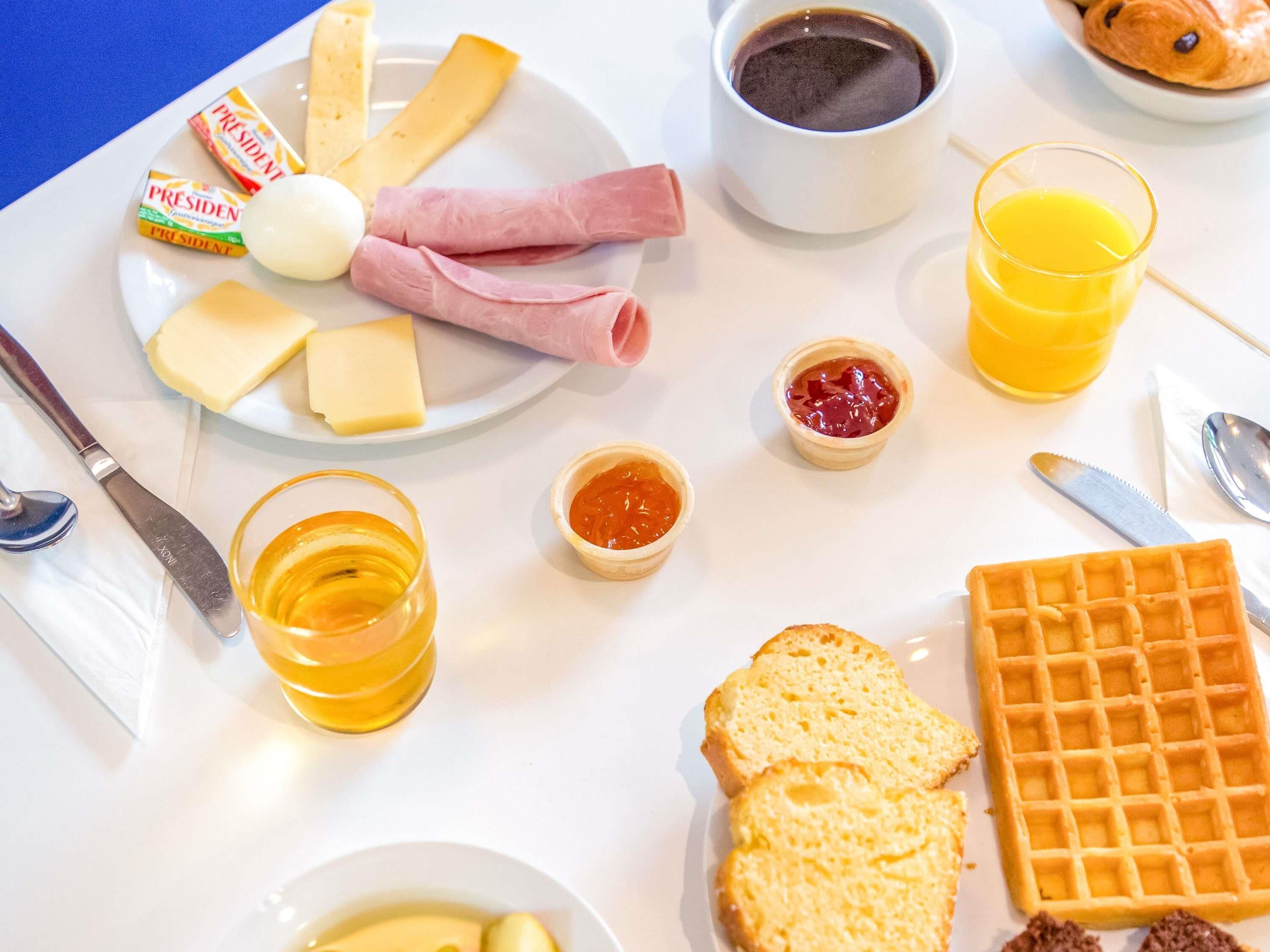 Une table de petit-déjeuner avec du fromage, du jambon, des œufs, de la confiture, du jus, du café, des toasts et une gaufre.
