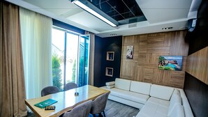 Een moderne hotelkamer met een houten eettafel, een witte bank en een groot raam met uitzicht op groen.