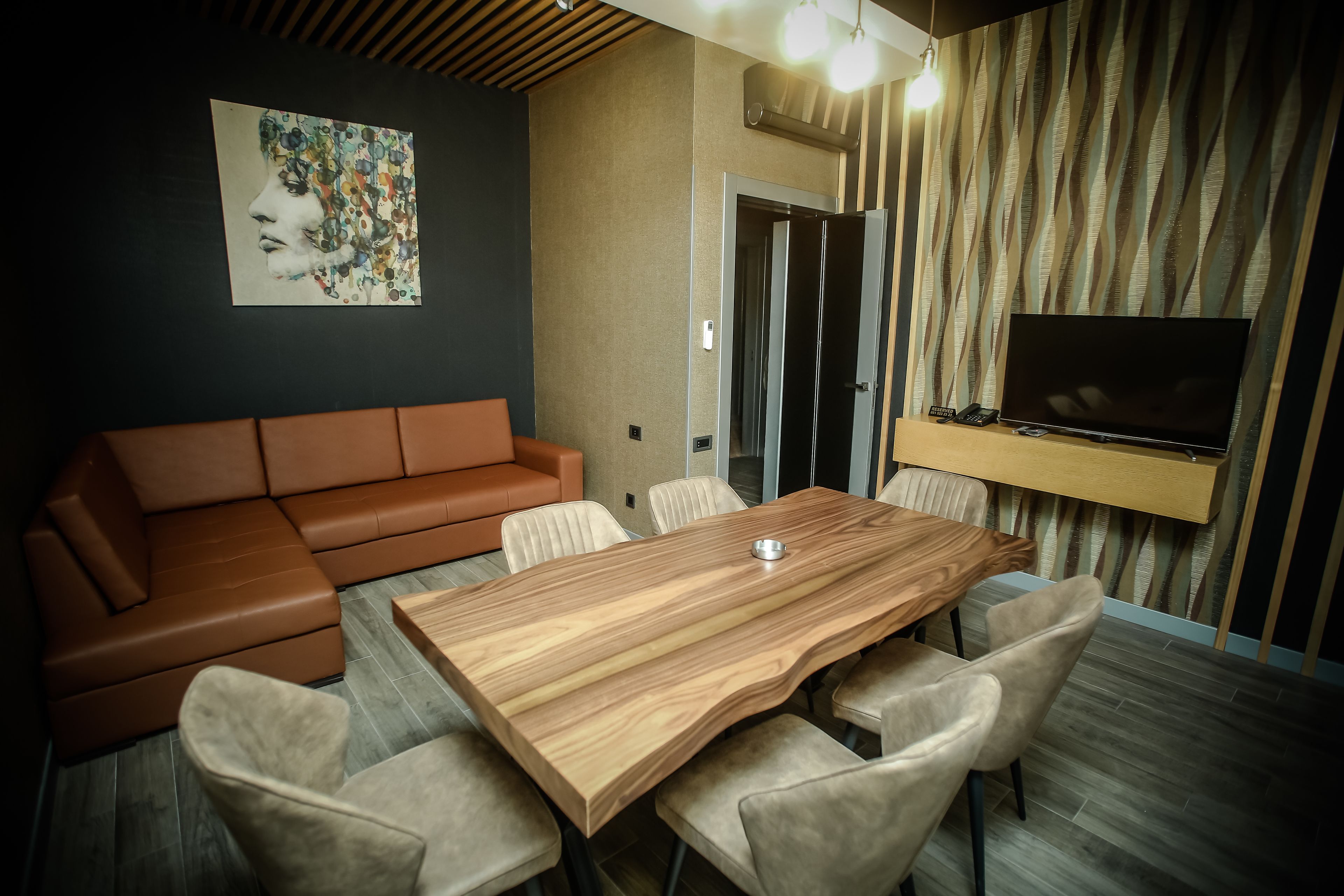 Una sala de estar moderna con una mesa de comedor de madera, sillas color beige, un sofá de cuero marrón, un televisor de pantalla plana montado en la pared y una cortina estampada.