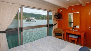 Ein Zimmer mit großem Fenster mit Blick auf einen See und ein Boot, ein Korbstuhl, ein Holztisch mit Spiegel und ein Bett mit blumigem Überwurf.