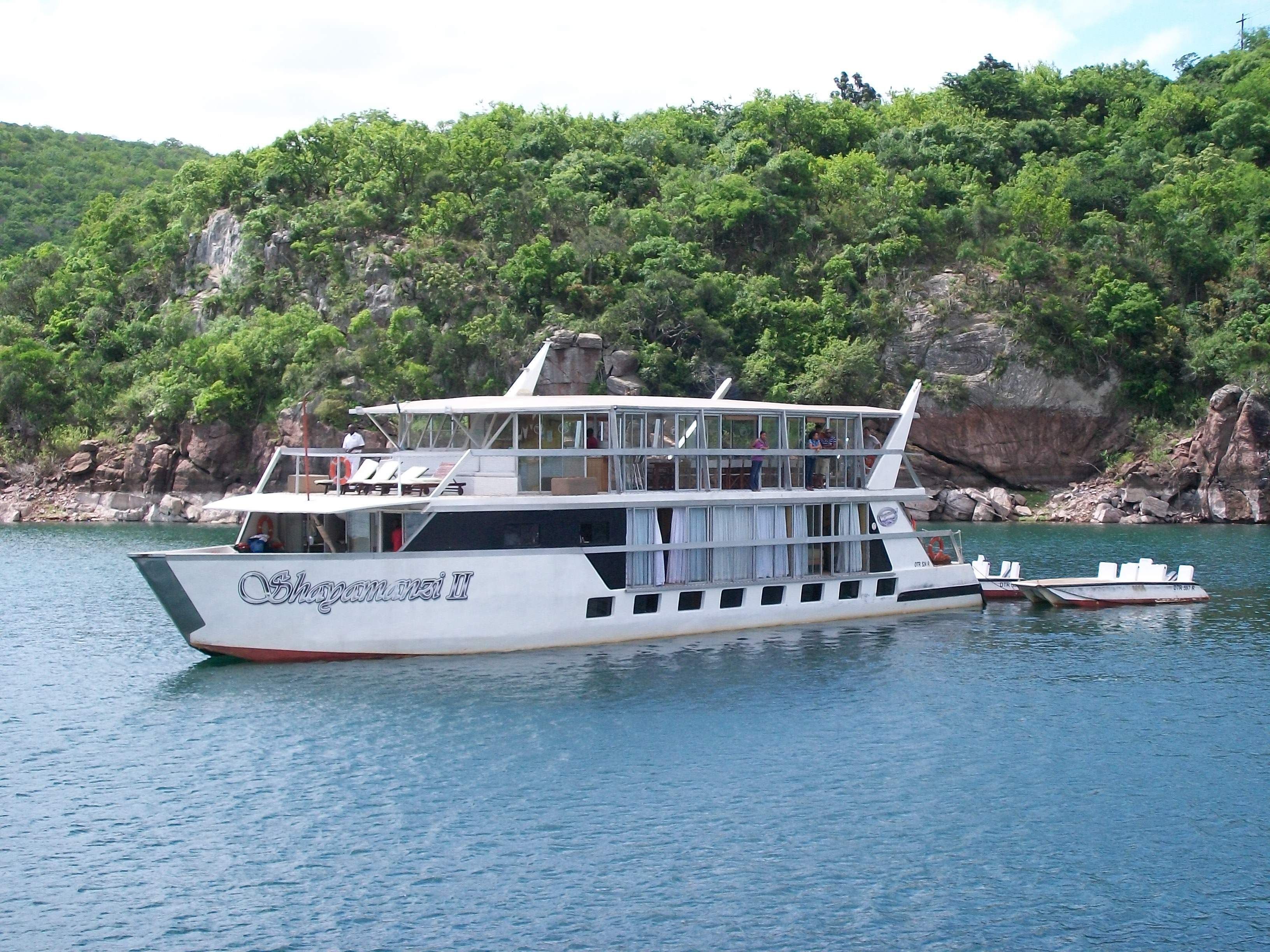 Un barco grande de varios pisos con un nombre visible en su costado, amarrado cerca de una costa rocosa con abundante vegetación.