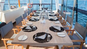 Ein Essbereich auf einem Kreuzfahrtschiff mit einem langen Tisch, der für eine Mahlzeit gedeckt ist, umgeben von Stühlen und großen Fenstern mit Blick auf das Meer.