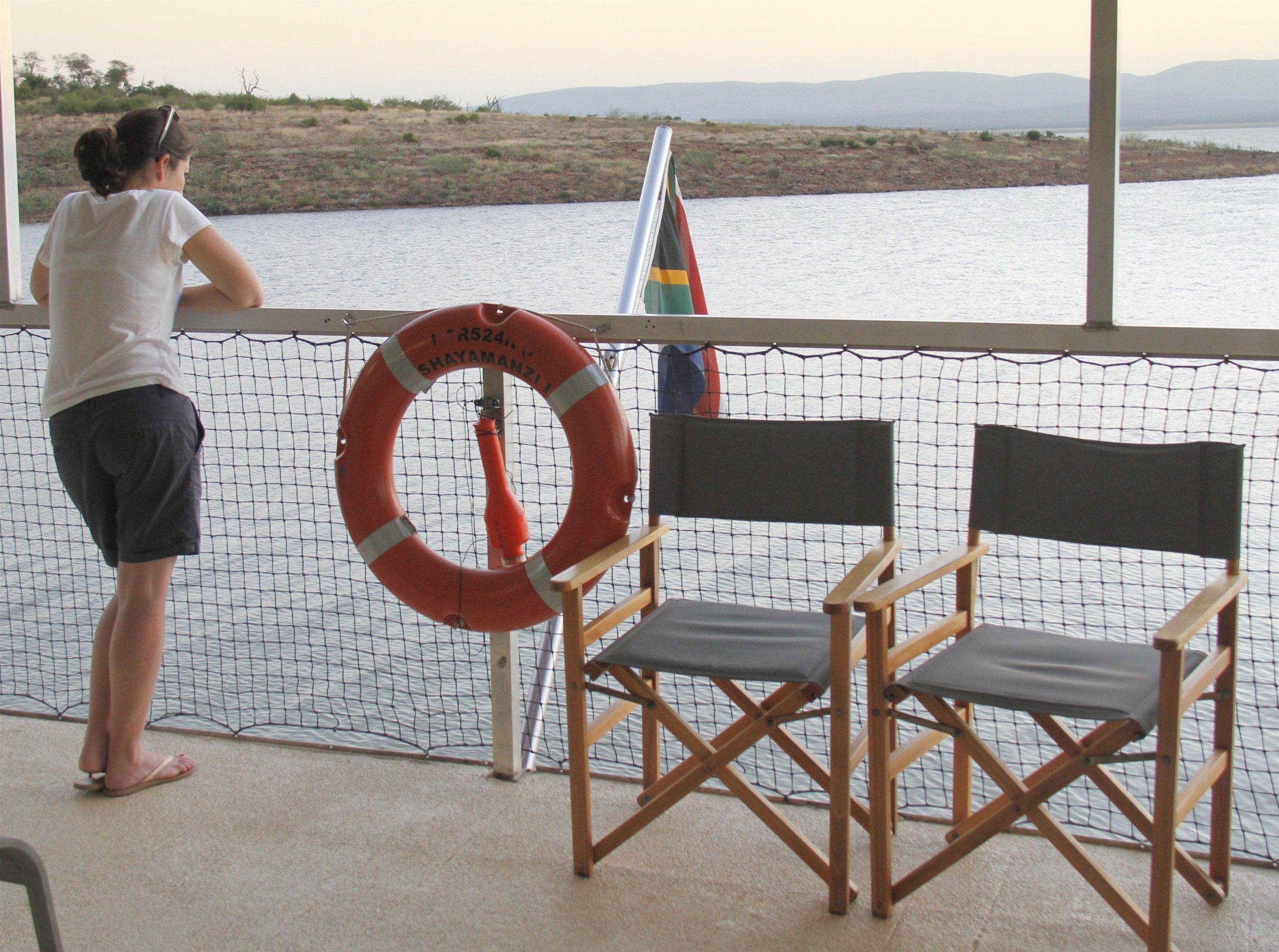 A person standing by a window overlooking a body of water, with a lifebuoy and a flag visible.