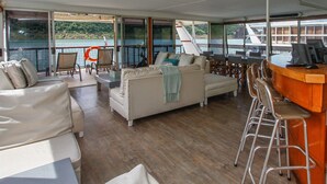 Eine großzügige Terrasse mit einer Sitzecke, Sesseln, einer Bar und großen Fenstern mit Blick auf ein Gewässer.