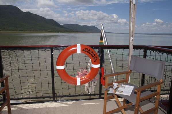 A lifebuoy attached to a boat's railing, a wooden chair, and a view of a calm lake with mountains in the background.