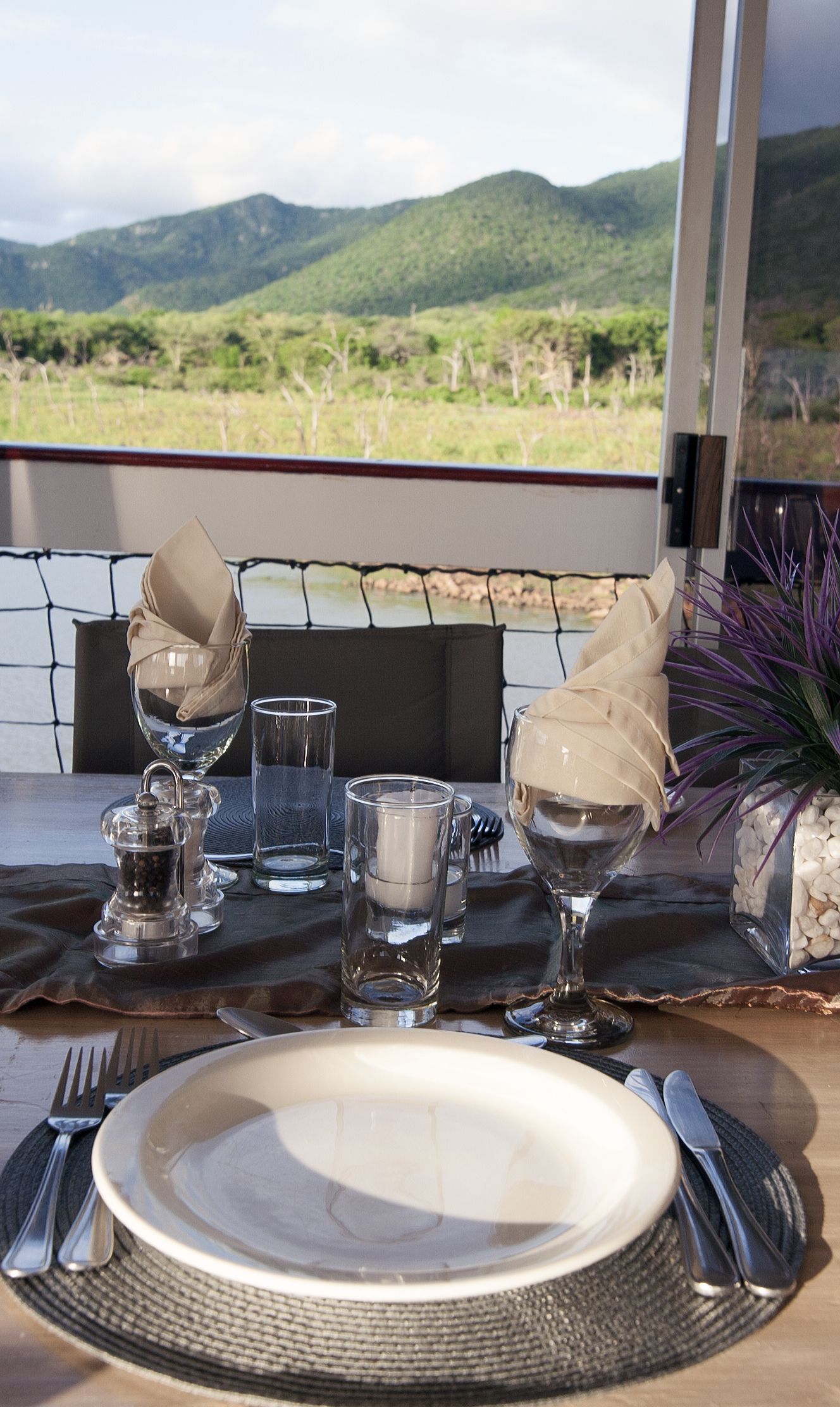 A dining table set with a white plate, cutlery, and clear glasses, with a scenic mountain view in the background.