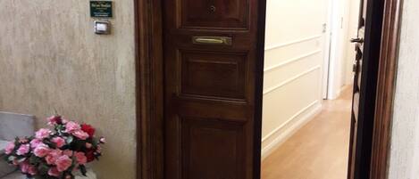 Una puerta de madera con una placa, un jarrón blanco con flores rosas y un felpudo estampado.