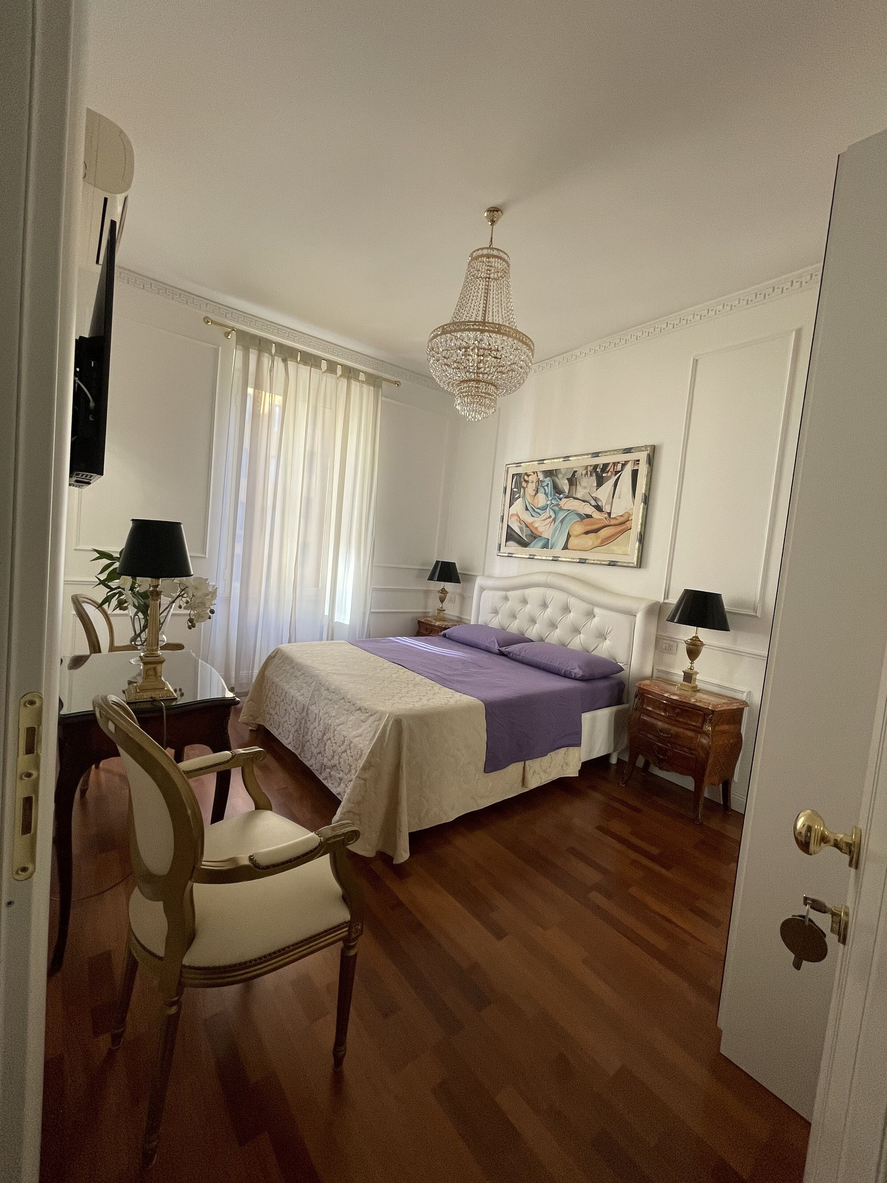 A bedroom with a bed, a chandelier, a painting, a desk, and a chair.