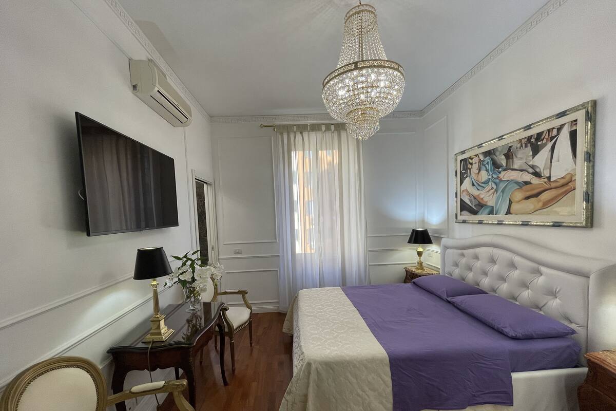 A hotel room with a large bed, a television, a chandelier, a painting, and a desk with a lamp.