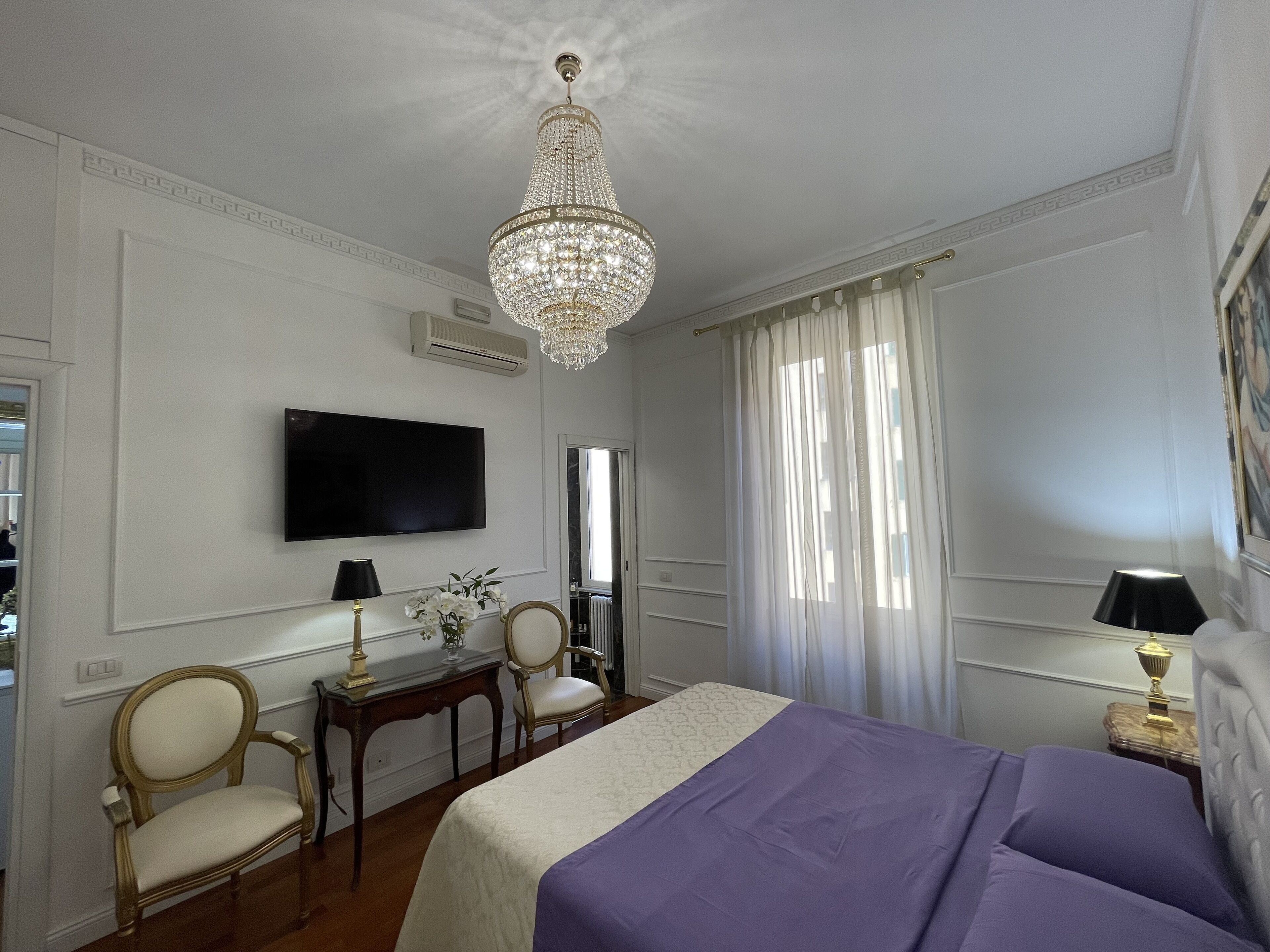 A hotel room with a large bed, a television, a chandelier, and two chairs.