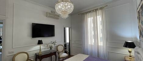 A hotel room with a large bed, a television, a chandelier, and two chairs.