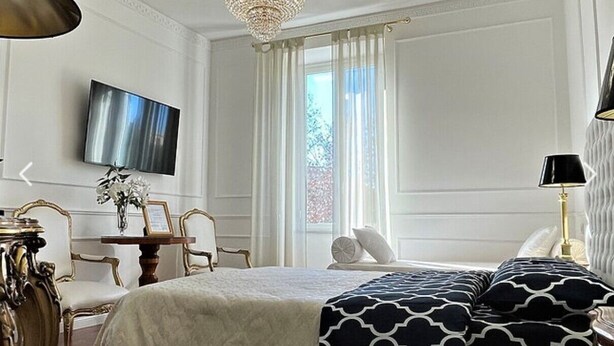 A hotel room with a bed, a TV mounted on the wall, a chandelier, a window with curtains, and a table with chairs. - King Plaza (Rome)