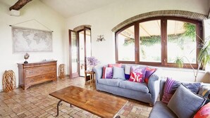 A living room with a wooden coffee table, a blue sofa with colorful cushions, a wooden dresser, and a large window with a view of greenery. - La Cavetta Country House (Trevignano Romano)