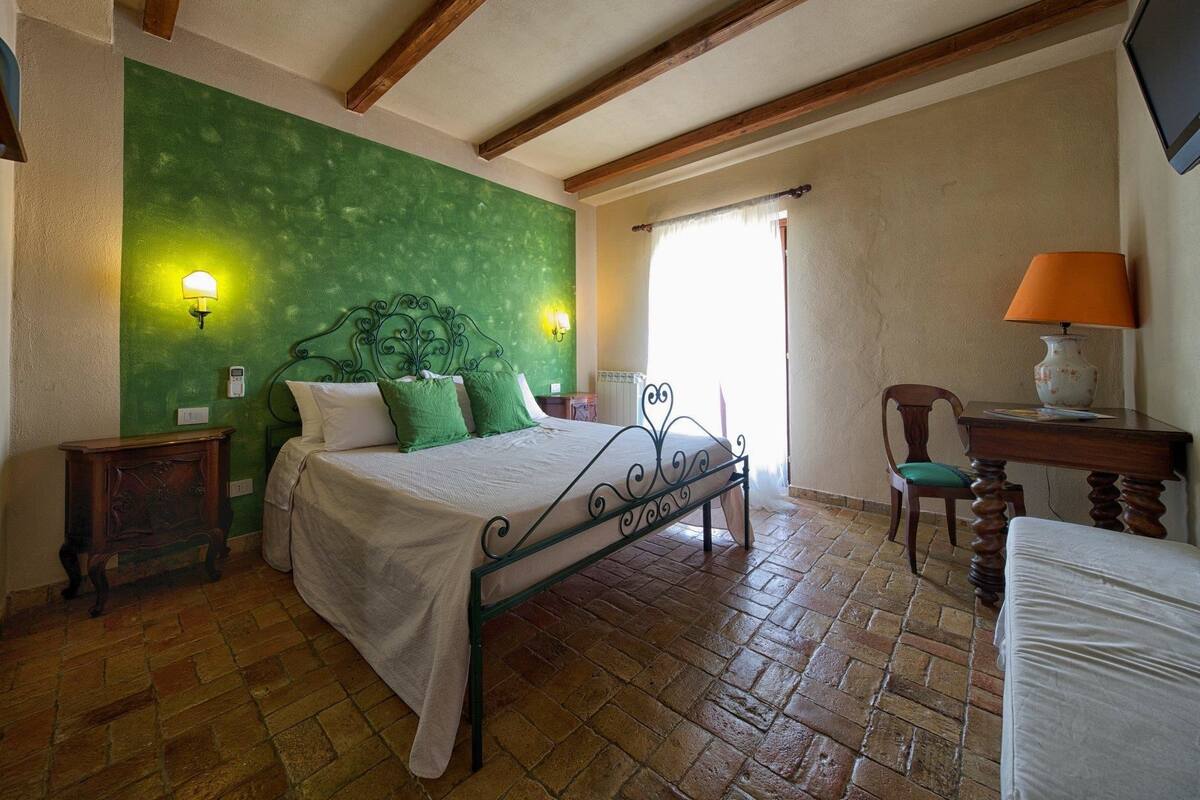 Een slaapkamer met een groene wand, een bed met metalen frame, een houten stoel, een bureau met lamp en een raam met gordijnen.