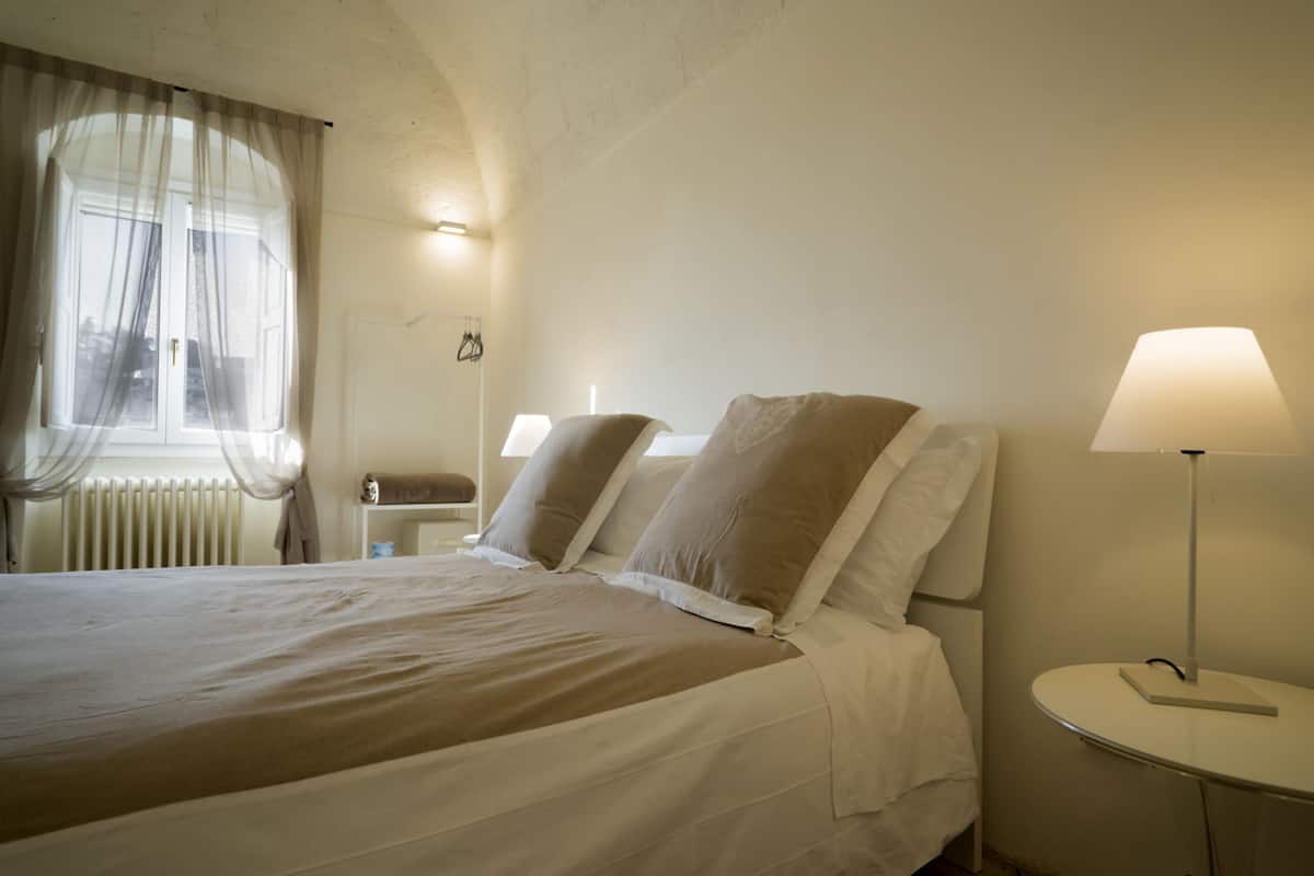 A neatly made bed with a white and beige bedspread, a bedside table with a lamp, and a window with sheer curtains.