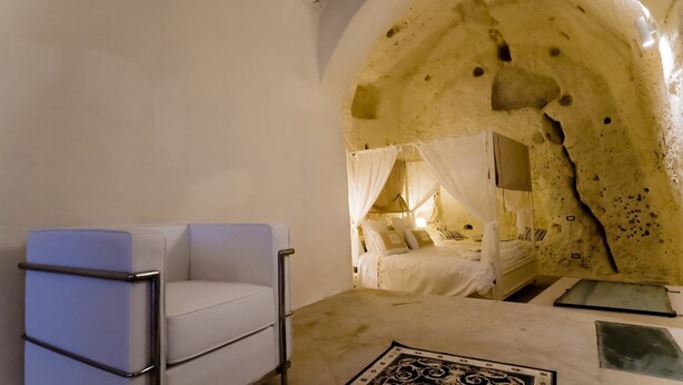 A room with a white cushioned chair, a bed with a canopy, and a unique cave-like interior. - Giardini di Pietra (Matera)