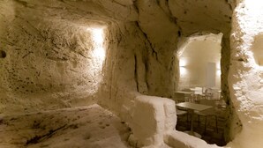 An interior with a rustic, cave-like stone wall, a narrow passage leading to a dining area with tables and chairs. - Giardini di Pietra (Matera)