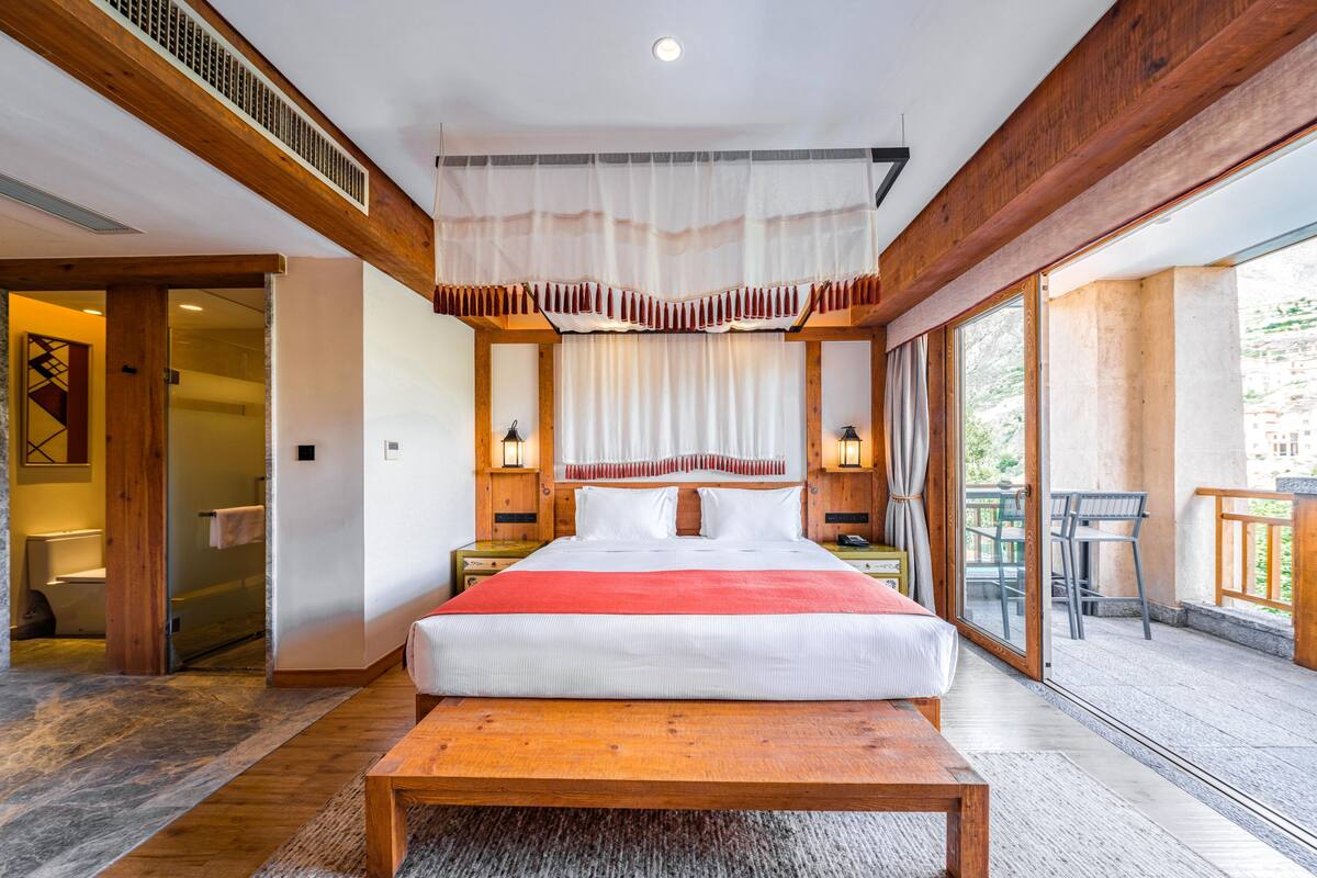 A hotel room with a large bed, wooden beams, a balcony with a table, and a view of the outside.