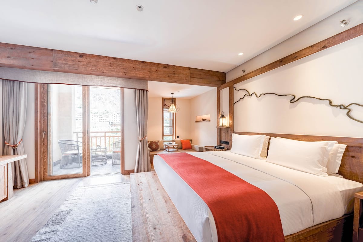 A bedroom with a large bed, wooden beams, a sliding glass door to a balcony, and a desk with a chair.