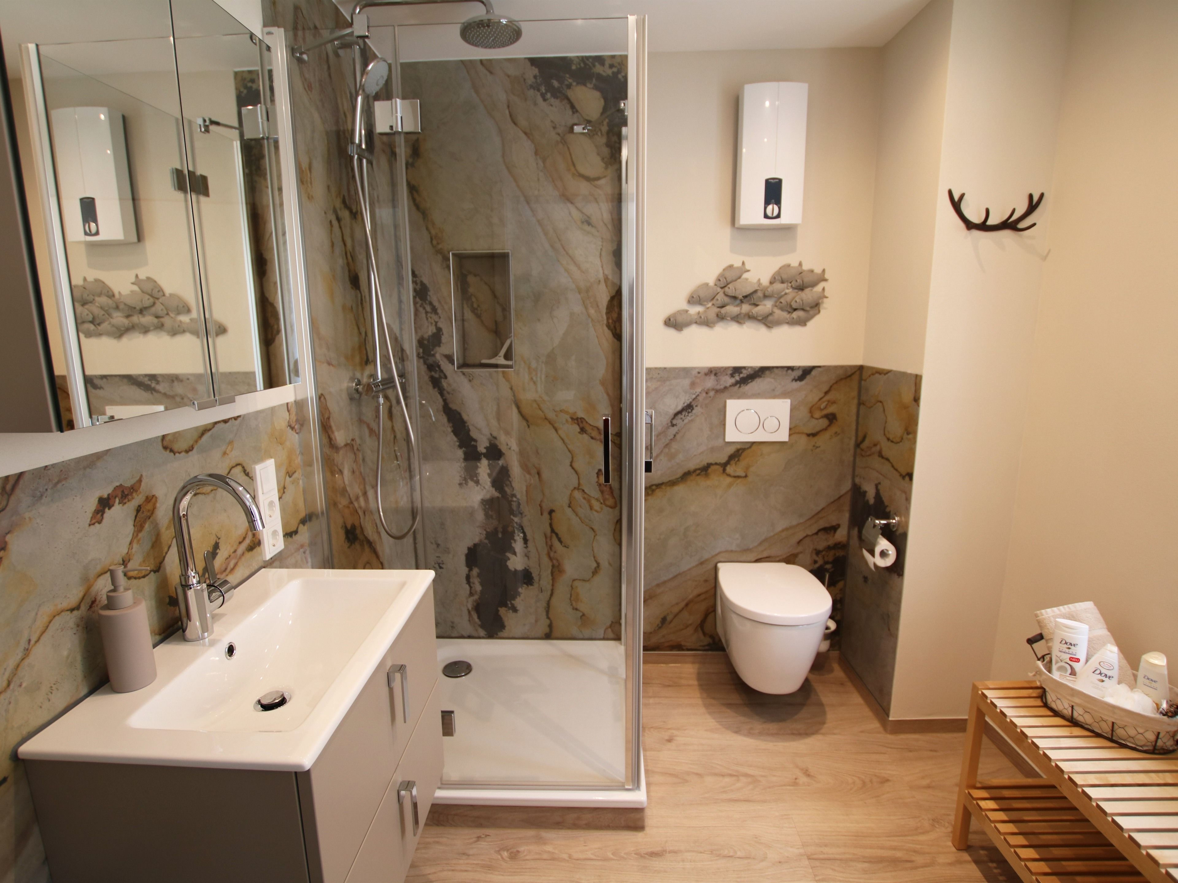 A modern bathroom with a glass-enclosed shower, a marble backsplash, a toilet, and a wooden bench with toiletries.