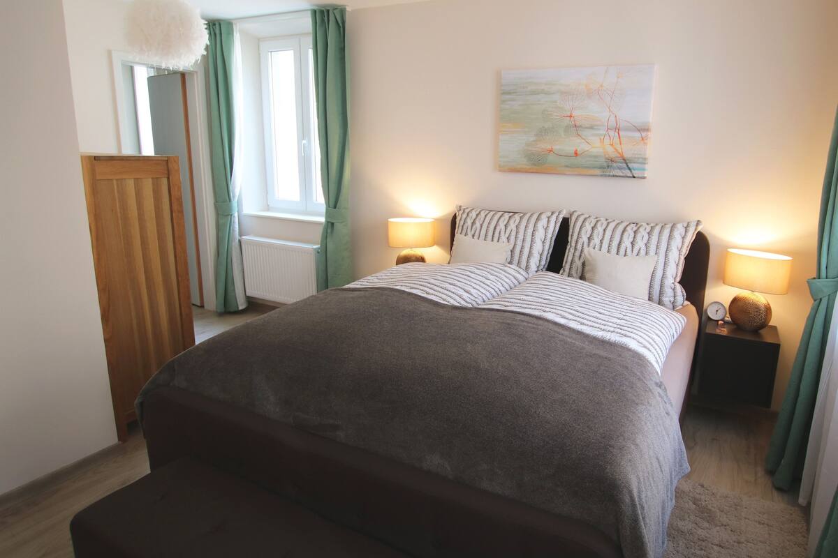Quarto com cama, duas mesas de cabeceira com luminárias, janela com cortinas, radiador e um quadro na parede.