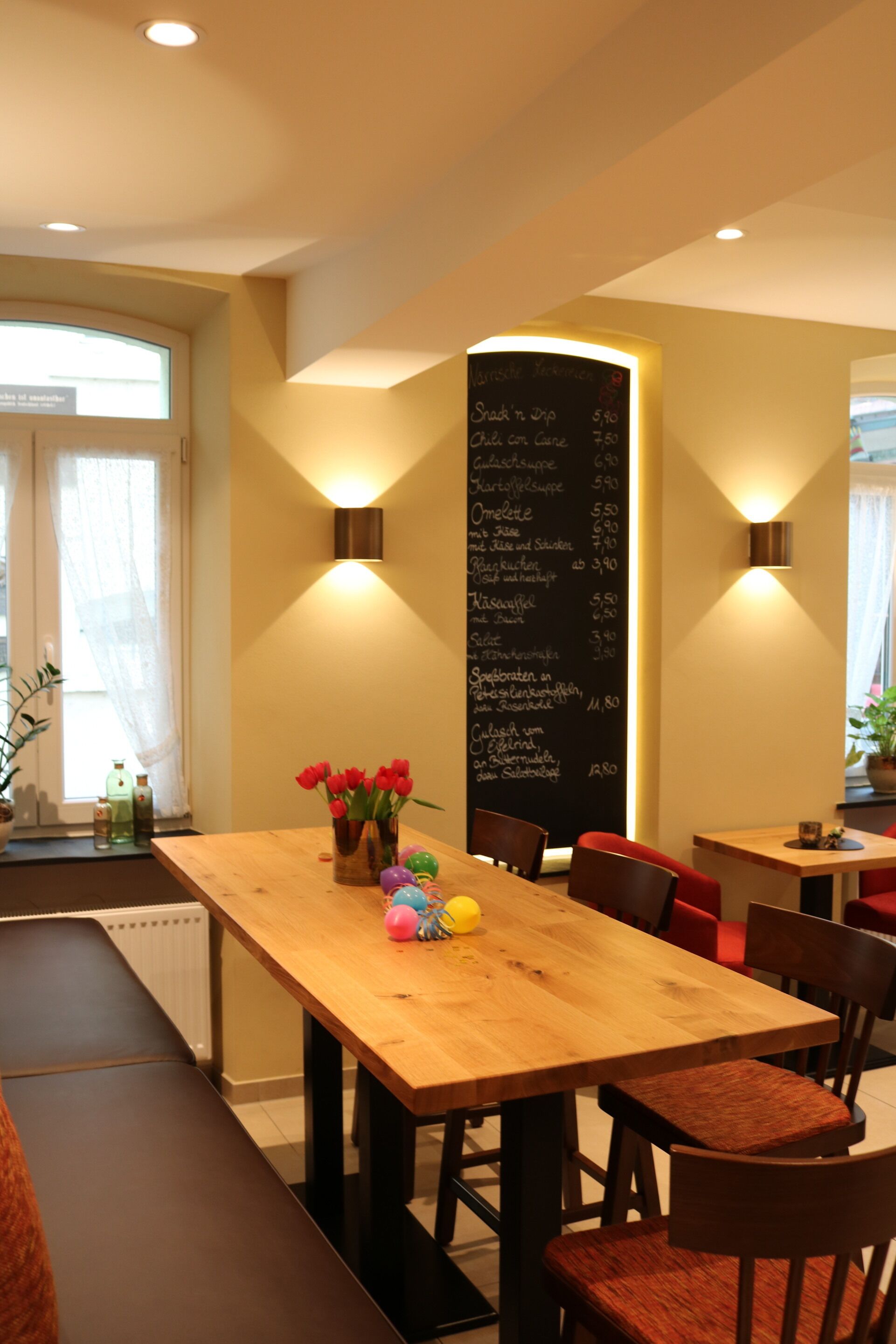 Un restaurant avec des tables et des chaises en bois, un menu écrit à la craie et un vase rempli de fleurs rouges.