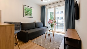 Un salon moderne avec un canapé gris foncé, une table basse en verre, une table d’appoint en bois et une grande fenêtre avec des rideaux.