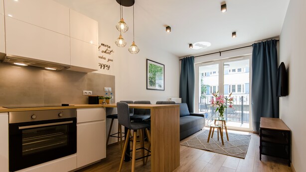 Une cuisine moderne dotée d’appareils électroménagers intégrés, un coin repas avec une table et des chaises en bois, et un espace de vie comprenant un canapé et de grandes fenêtres.