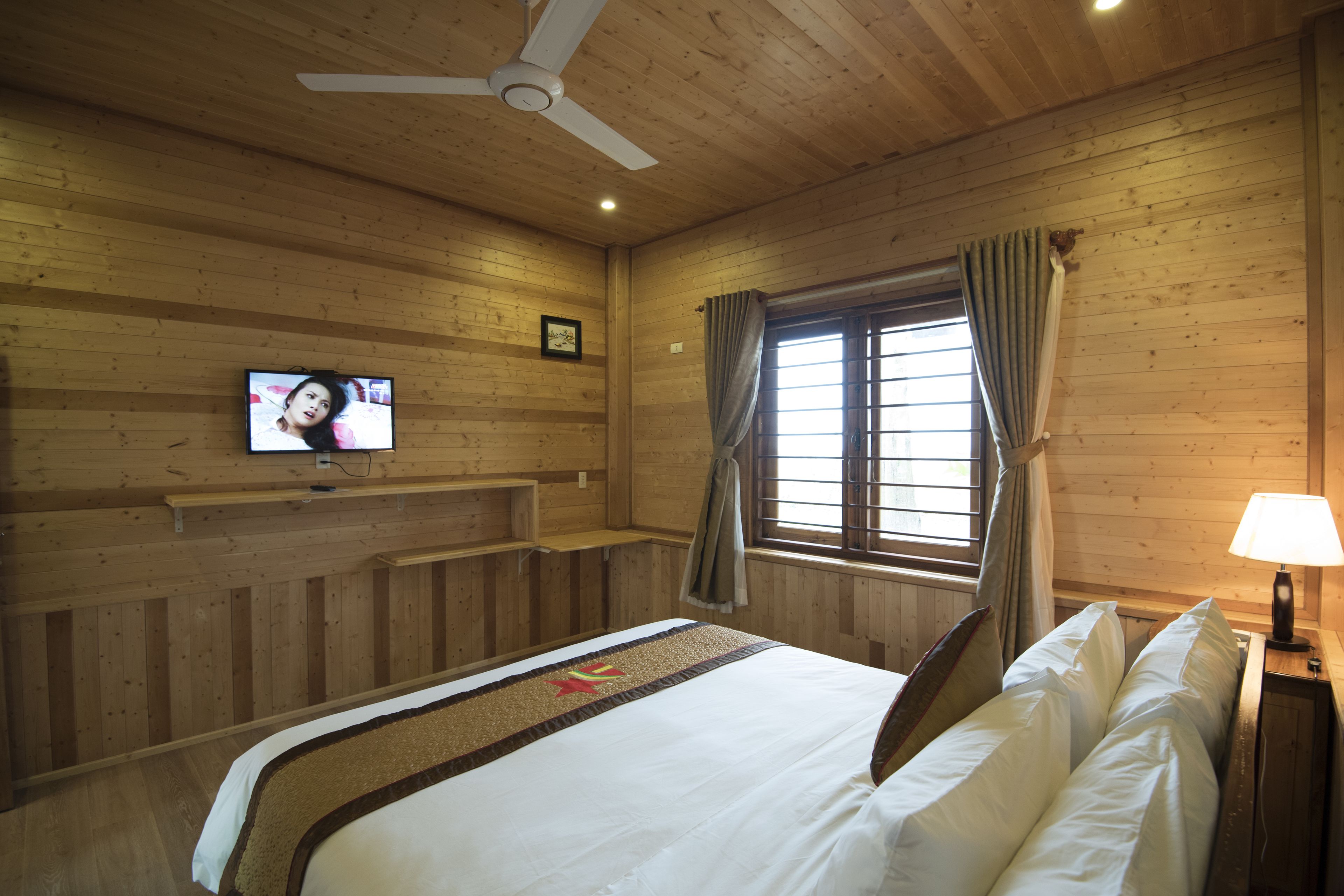 Una habitación con paneles de madera, una cama, un televisor, una ventana con cortinas y un ventilador de techo.