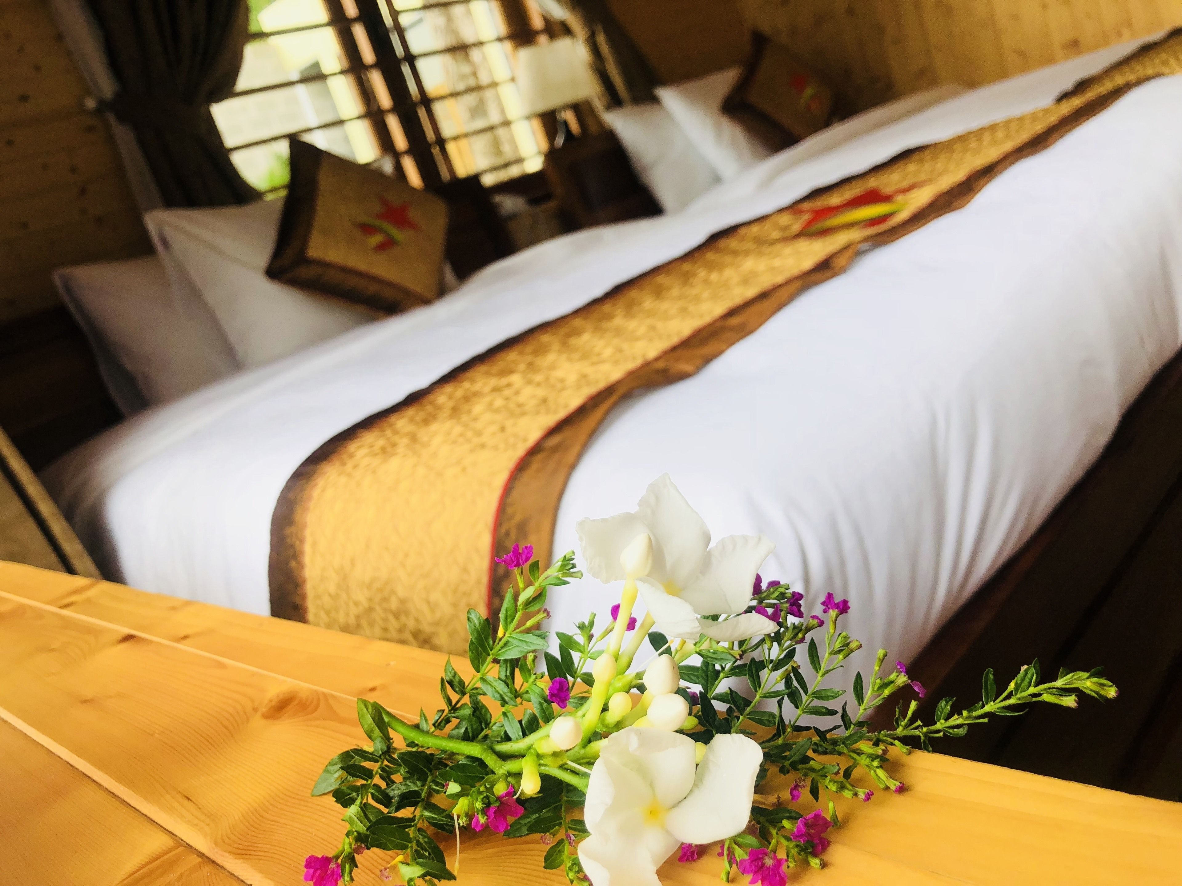 A neatly made bed with white linens and a brown decorative strip, a wooden table with a floral arrangement, and a window with curtains in a room.